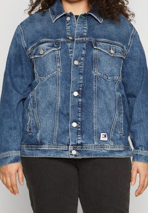 Femme portant une veste en jean bleue à boutons avec poches poitrine et pantalon marron, mains détendues le long du corps, torse visible du cou aux hanches.