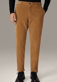 Man met een beige corduroy broek met opgerolde broekspijpen en zwarte schoenen, staande met één hand in zijn zak.