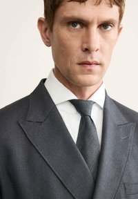 Completo grigio su misura con rever a puntino, accompagnato da una camicia bianca e una cravatta grigia texturizzata. Primo piano sul corpo superiore.