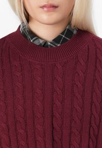 Burgunderfarbener Strickpullover mit Zopfmuster, geripptem Rundhalsausschnitt, über einem Schwarz-Weiß-karierten Hemd getragen.