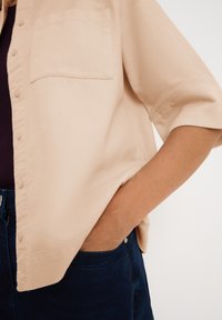 Camisa de manga corta beige con tejido texturizado, un bolsillo delantero y botones, llevada sobre una blusa oscura, combinada con pantalones azul oscuro.
