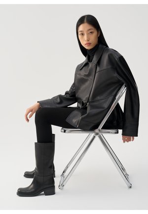 Młoda kobieta z prostymi czarnymi włosami, ubrana w czarną skórzaną kurtkę, prążkowane spodnie i buty, siedzi bokiem na chromowanym składanym krześle.
