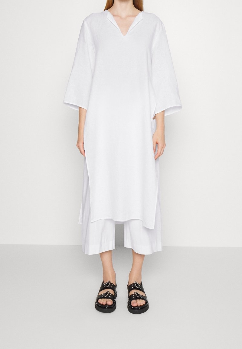 Personne vêtue d'une longue tunique blanche ample avec des fentes latérales, d'un pantalon blanc large et court, et de sandales noires à boucle, debout devant un fond blanc uni.