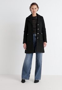 Abrigo de lana negro con cuello en pico, botones plateados y bolsillos laterales, combinado con un suéter oscuro texturizado y jeans de pierna ancha azul.