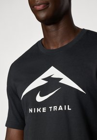 Schwarzes T-Shirt mit Rundhalsausschnitt. Mit einem großen weißen Berggrafik und dem Text "NIKE TRAIL" in fetten weißen Buchstaben. Aus Baumwollmaterial.
