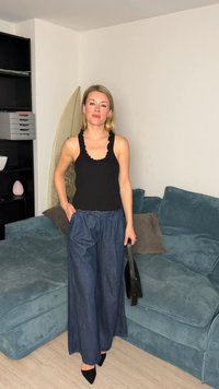 Kobieta stojąca w salonie, ubrana w czarną bluzkę bez rękawów, szerokie niebieskie spodnie, czarne buty, trzymająca czarną torebkę obok turkusowej sofy i deski surfingowej.