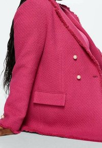Femme portant un blazer rose vif texturé avec des boutons en perle, assise avec un bras reposant sur le sol, montrant de longs cheveux foncés.