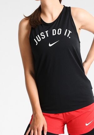 Frau trägt ein schwarzes ärmelloses Nike-Shirt mit dem Schriftzug „JUST DO IT.“ und rote Nike-Sportshorts, steht mit einer Hand in der Hüfte.