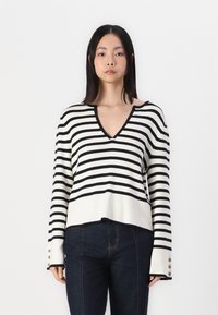 Black and white striped sweater with a V-neck, ribbed texture, and buttoned cuffs. Worn with dark denim jeans.