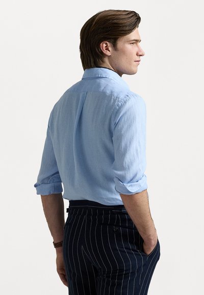 Világoskék, puha anyagból készült ing, felgöngyölt ujjak és laza fazon jellemzi, ami sötétkék csíkos nadrággal lett párosítva.