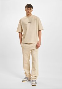 Béžové volné bavlněné tričko s malým černým logem, spojené se sladěnými lehkými béžovými kalhotami a šedo-bílými teniskami.