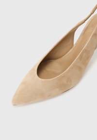 Zapato de tacón tipo slingback de ante beige con puntera puntiaguda y diseño de lado recortado, que presenta un forro interior suave y un tacón delgado.