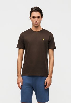 Muž oblečený v jednoduchom tmavohnedom tričku s krátkym rukávom a malým žltým logom, oblečený v modrých kraťasoch po kolená, stojaci na bielom pozadí.