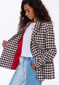 Žena s kudrnatými vlasy, oblečená v černozeleném a růžovém kostkovaném saku, které ukazuje červenou podšívku, bílou košili a dvoubarevné modré džíny, s jednou rukou v kapse.