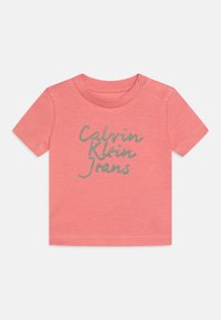 Coral cotton T-shirt with short sleeves and a round neckline. Features silver glitter text "Calvin Klein Jeans" across the front.