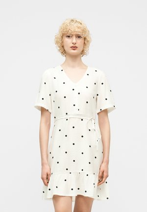 Persoană blondă purtând o rochie scurtă albă cu buline negre, decolteu în V, mâneci scurte și tiv cu volane, stând în picioare pe un fundal simplu.