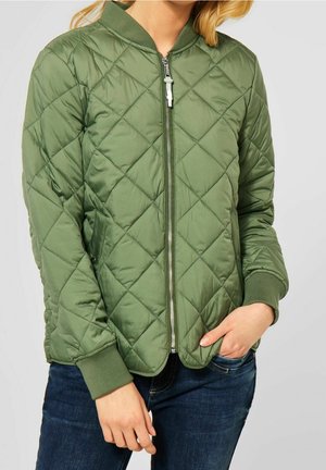 Vrouw draagt een olijfgroene doorgestikte bomberjas met een rits aan de voorkant en geribde manchetten, gecombineerd met donkerblauwe jeans.