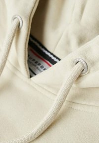 Beige hoodie collar with silver eyelets and beige drawstrings. Features a black and red label with text, showing fabric texture and stitching details.