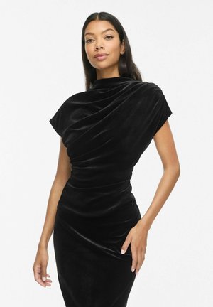 Femme aux longs cheveux foncés portant une robe en velours noir moulante avec des détails drapés et un col montant, posant devant un fond blanc uni.