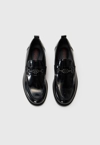 Mocasines de charol negro con una textura suave, que presentan una correa decorativa y un acento metálico, puntera redondeada y plantilla acolchada.