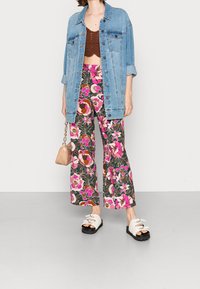 Džínová bunda přes hnědý crop top, květinové široké kalhoty v růžové a zelené barvě a bílé sandály s černými podrážkami. Drží malou béžovou kabelku.