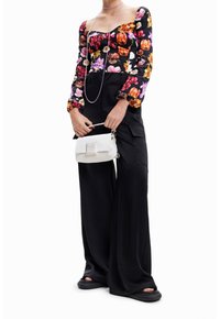 Černý top s květinovým vzorem a dlouhými rukávy, ve spojení s širokými černými kalhotami. Kompaktní bílá kabelka s logem v přední části. Černá obuv s otevřenou špičkou.