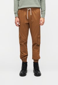 Καφέ παντελόνι εργασίας με ελαστικές μανσέτες, πλαϊνές τσέπες και ζωνάκι με κορδόνι στη μέση. Το υλικό φαίνεται λείο με ματ φινίρισμα. Φορεμένο με μαύρες μπότες.