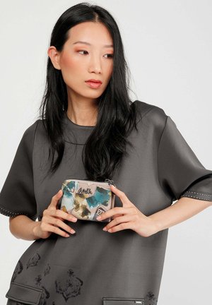 Bolsa con estampado floral con acentos en azul, verde y beige; cuenta con cierre de cremallera y un logo metálico en la parte delantera, sostenida por una persona.