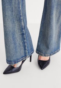 Des chaussures à talons hauts en cuir noir avec un bout pointu, portées avec un jean en denim bleu à jambes larges présentant des ourlets effilochés et un léger délavage.