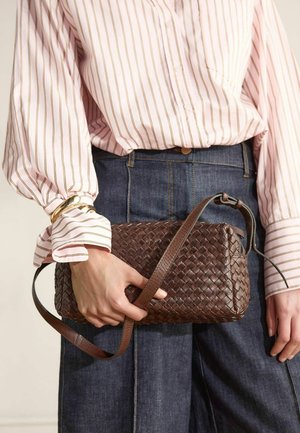 Persoon die een roze gestreepte blouse en donkere spijkerbroek draagt, houdt een gevlochten bruine leren handtas met een verstelbare riem vast.