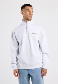 Felpa grigia chiaro con collo a zip fino a un quarto, con "New York" ricamato in verde sul petto. Tessuto morbido, vestibilità comoda.