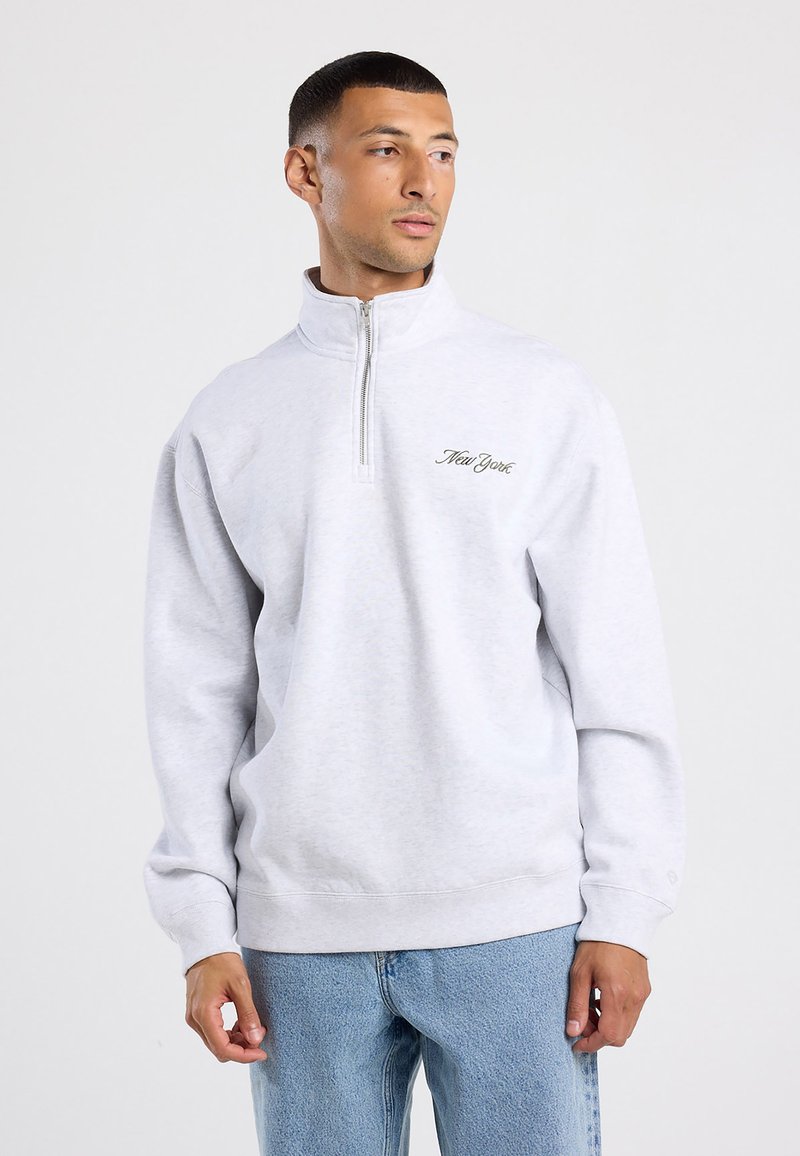 Felpa grigia chiaro con collo a zip fino a un quarto, con "New York" ricamato in verde sul petto. Tessuto morbido, vestibilità comoda.