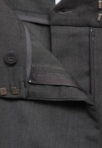 Schwarze Jeans halten mit einem Knopf- und Reißverschluss, sichtbaren Nähten und Metallakzenten, die eine strukturierte Stoffoberfläche zur Schau stellen.