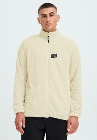 Cremefarbene Fleecejacke mit hohem Kragen, Frontreißverschluss und einem Labelpatch auf der linken Brust. Weicher Stoff und lockere Passform.