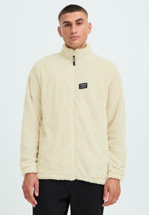 Cremefarbene Fleecejacke mit hohem Kragen, Frontreißverschluss und einem Labelpatch auf der linken Brust. Weicher Stoff und lockere Passform.