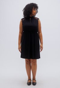 Μαύρο φόρεμα με ψηλό λαιμό, χωρίς μανίκια και δαντελωτές λεπτομέρειες στους ώμους. Αέρινο ύφασμα με ελαστική μέση, συνοδευόμενο από μαύρα παπούτσια.