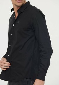 Chemise noire à manches longues et boutons, avec un col pointu, des boutons blancs et un petit logo sur la poitrine. Tissu lisse et coupe ajustée.