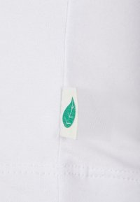 Vitt bomullstyg med en grön bladlogotyp på sidan. Texturen är slät, och sömmarna är synliga längs fållen.