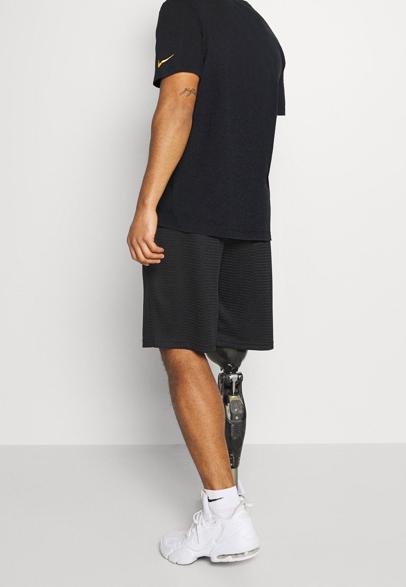 Czarna koszulka Nike z żółtym logo, czarne spodenki o teksturowanym wzorze i białe sneakersy. Widoczna proteza nogi z metalicznymi i teksturowanymi detalami.