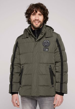 Man smiling wearing olive green padded jacket with black stripes on sleeves and patch logos on chest and sleeve, standing against plain background.