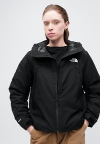 Černá nepromokavá bunda s kapucí, dvěmi kapsami na zip a nastavitelnými manžetami. Na levé části hrudi se nachází logo a bunda má hladkou texturu.