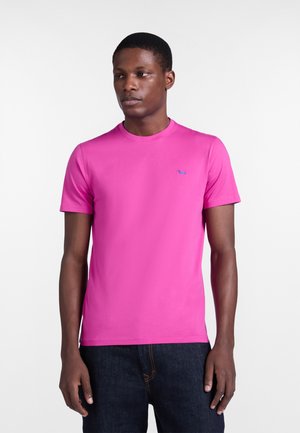 Muž oblečený v jasne ružovom tričku s krátkym rukávom a malým modrým logom psa, v kombinácii s tmavomodrými denimovými džínsami, stojaci pred jednoduchým bielym pozadím.