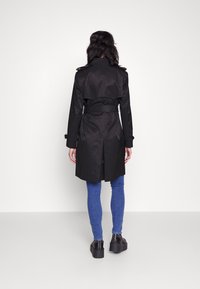 Svart trench coat med bälte i midjan, dubbelknäppt design och axelklädslar, i kombination med blå smala jeans och svarta platåstövlar.