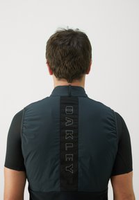 Čierna a tmavomodrá cyklistická vesta s zipsovým golierom, ktorá má na zadnej strane výrazné vertikálne logo "OAKLEY" a textúrované rukávy.