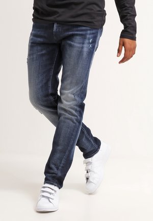 Homme portant un jean bleu foncé et des baskets blanches, debout jambes croisées devant un fond uni.