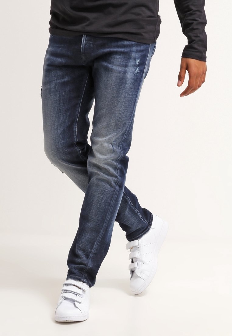 Homme portant un jean bleu foncé et des baskets blanches, debout jambes croisées devant un fond uni.