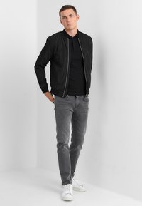 Giubbotto bomber nero con polsini a costine, abbinato a una maglietta polo nera, jeans grigi slim-fit e sneaker bianche. Sfondo neutro.