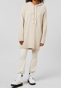 Beige gebreide trui met capuchon en trekkoorden, geribbelde boorden en zoom. Gecombineerd met lichtbeige taps toelopende broek en witte schoenen.