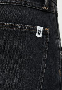 Czarna kieszeń z dżinsu z pomarańczowym przeszyciem i małą białą metką z logo czarnego chrząszcza w prawym górnym rogu.