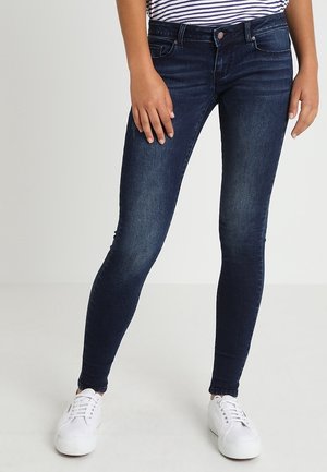 Personne portant un jean skinny bleu foncé, des baskets blanches et une chemise rayée, debout les jambes légèrement écartées sur un fond uni.